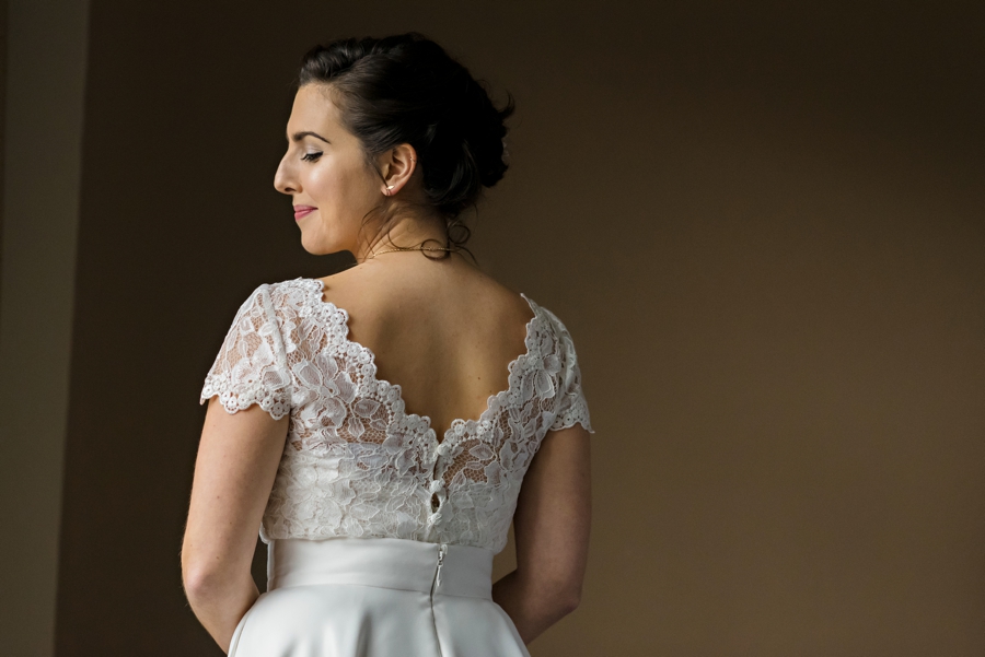 A portrait of a bride in an open back lace wedding dress next to a window.